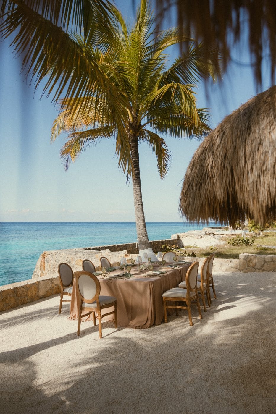 A long table in white linen under the palapa, the sea behind it